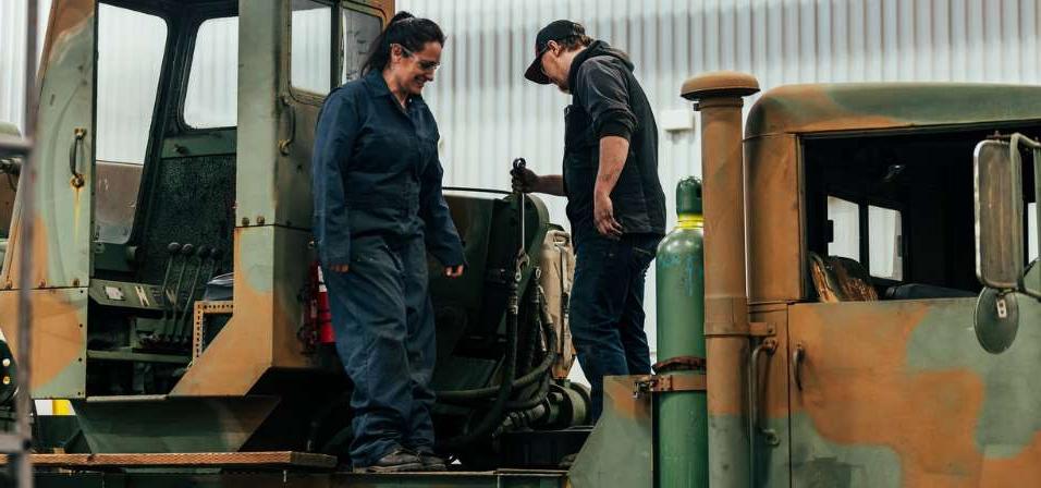 一男一女学生站在一辆军用彩绘卡车上笑着，其中一个拿着扳手准备拧紧软管